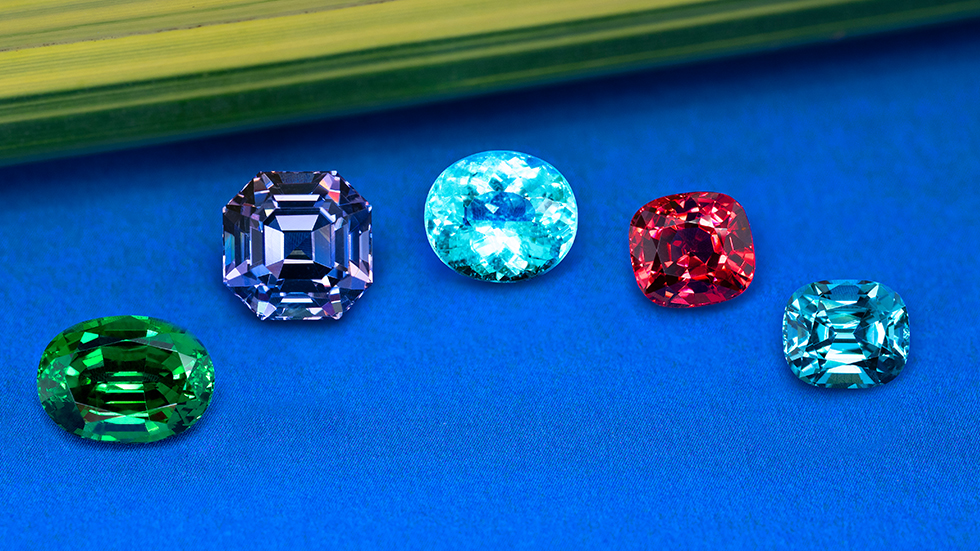 Side-by-side individual gemstones on a blue background: oval deep forest green tsavorite garnet, radiant cut blue purple tanzanite, oval neon electric blue paraiba tourmaline, intense stoplight red spinel cushion-cut, and blue indicolite tourmaline cushion-cut.