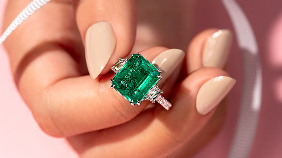 A photo of a 5 carat Emerald ring which has a Diamond on each side, held gently towards the camera.
