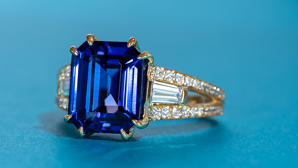 Three-stone deep blue tanzanite ring with diamond baguettes and a pave band in yellow gold.