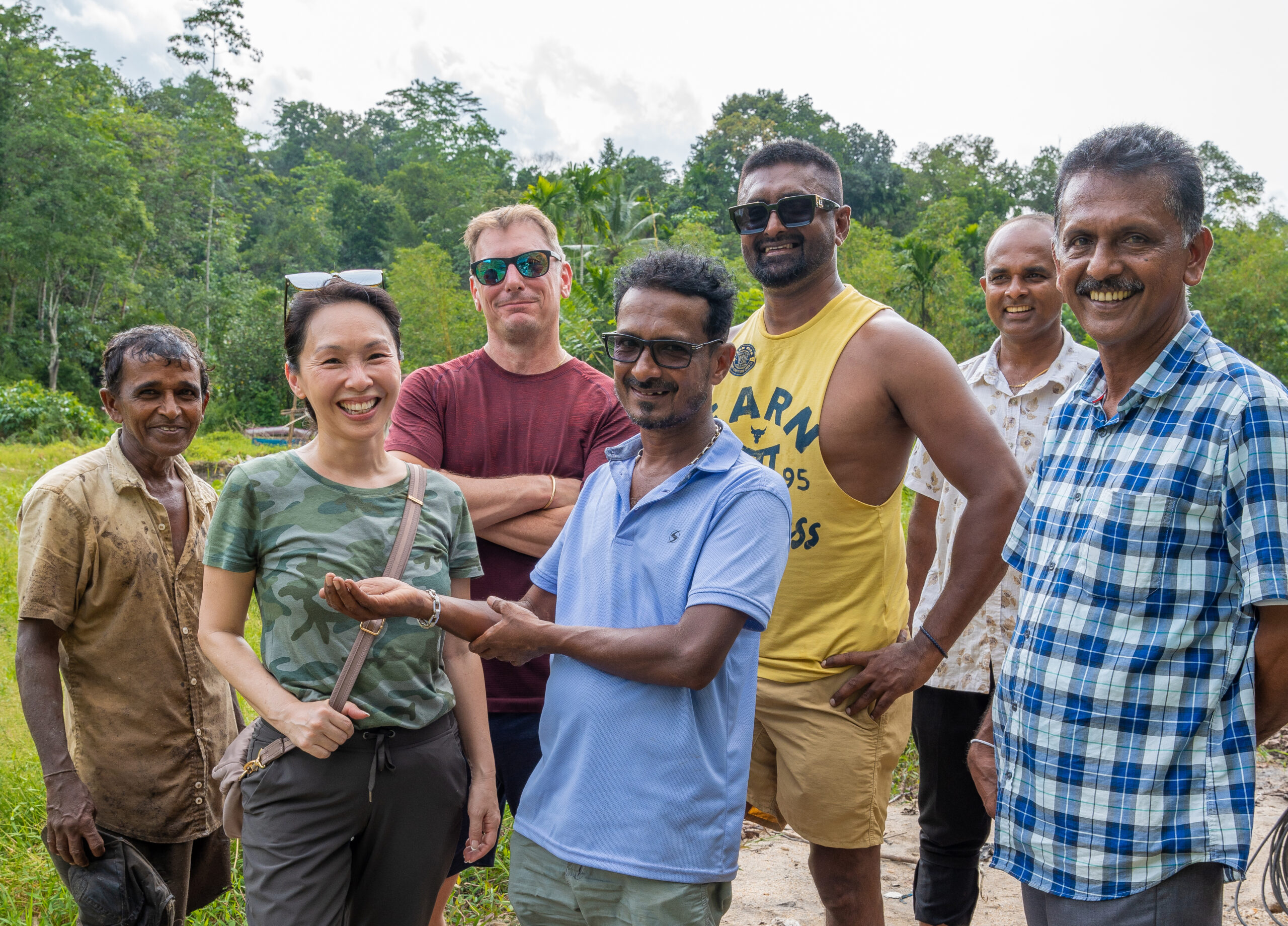 A diverse group of individuals, including a Chinese woman and a Caucasian, standing among joyful Sri Lankan men at a Sri Lanka mining site, all sharing smiles
