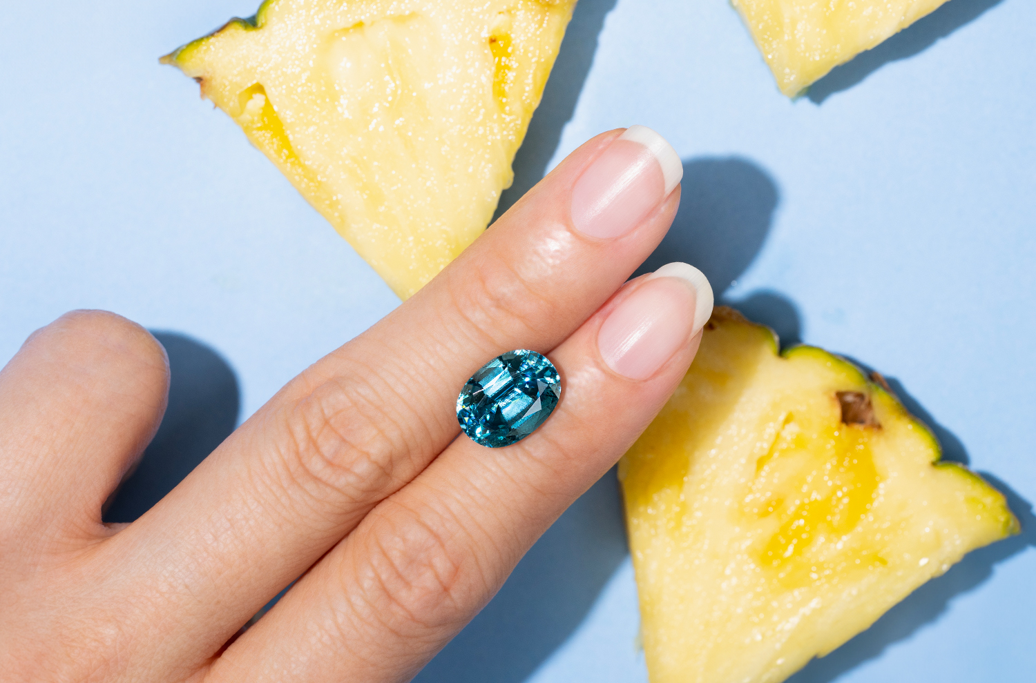 A fun, summer themed photo of an oval-shaped blue Zircon with a vibrant background of blue hues and bright yellow pineapple slices.