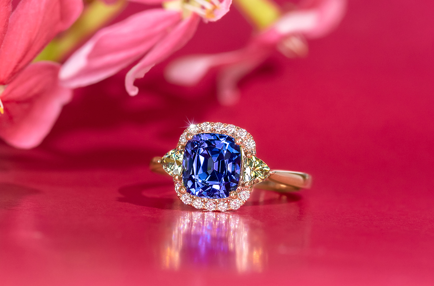 A ring featuring a vibrant and deep blue Tanzanite as its main gemstone. The tanzanite is surrounded by a classic diamond halo and flanked by smaller, trillion cut Garnets.